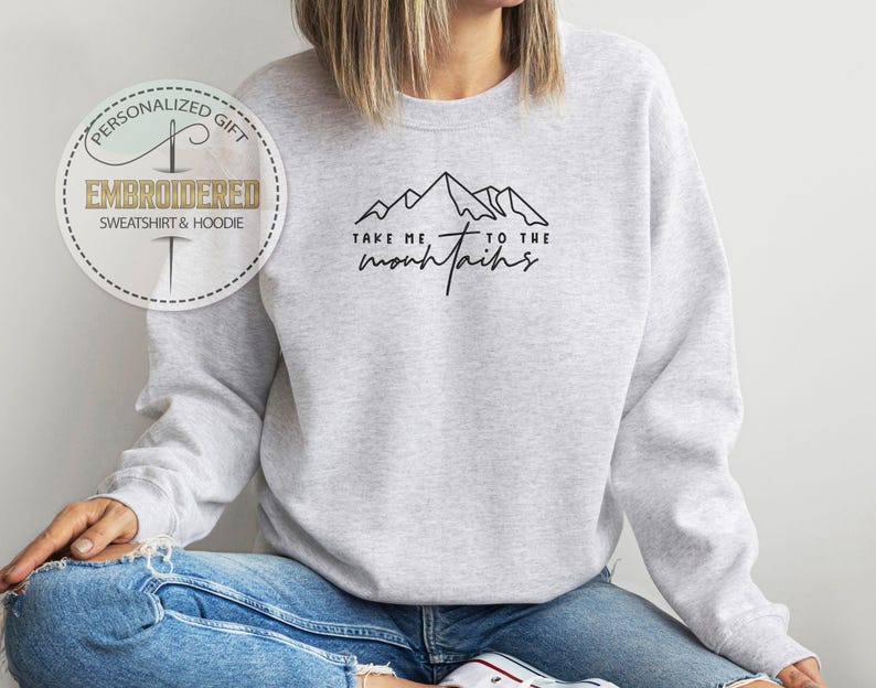 K&ouml;nnte beinhalten: Hellgraues Sweatshirt mit dem schwarzen Schriftzug "Take me to the mountains" und einer Berg-Grafik. Das Sweatshirt hat auch ein rundes Logo mit der Aufschrift "Personalized Gift, Embroidered Sweatshirt & Hoodie".