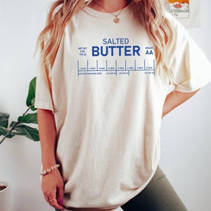 May include: A cream-coloured t-shirt featuring the words "SALTED BUTTER" in blue, with a measurement guide. The shirt also states the net weight as 113 g and the grade as AA. The t-shirt is a casual style.