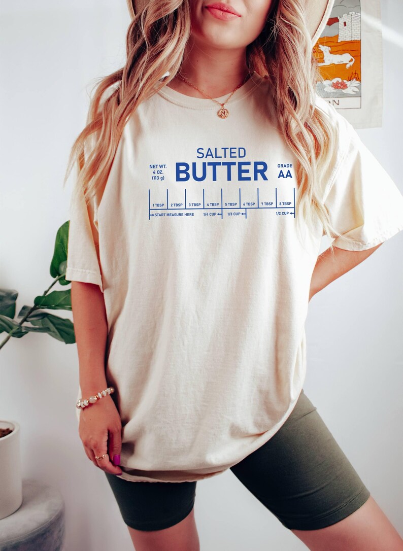 May include: A cream-coloured t-shirt featuring the words "SALTED BUTTER" in blue, with a measurement guide. The shirt also states the net weight as 113 g and the grade as AA. The t-shirt is a casual style.