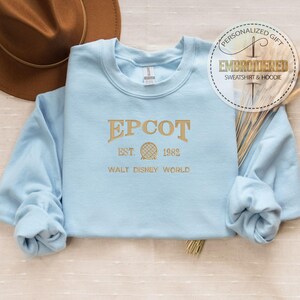 May include: A pale blue sweatshirt with gold embroidered text reading "EPCOT EST. 1982 WALT DISNEY WORLD". A brown hat is in the background. The sweatshirt has long sleeves and a crew neck. A circular sticker reads "PERSONALIZED GIFT EMBROIDERED SWEATSHIRT & HOODIE".