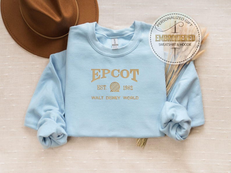May include: A pale blue sweatshirt with gold embroidered text reading "EPCOT EST. 1982 WALT DISNEY WORLD". A brown hat is in the background. The sweatshirt has long sleeves and a crew neck. A circular sticker reads "PERSONALIZED GIFT EMBROIDERED SWEATSHIRT & HOODIE".