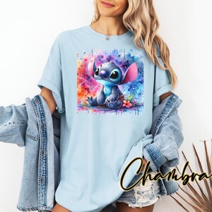 May include: Pale blue t-shirt with a watercolour illustration of the Disney character Stitch. The artwork depicts Stitch in a seated position against a backdrop of pink, blue, and purple colours. The t-shirt is made of a soft, comfortable material.