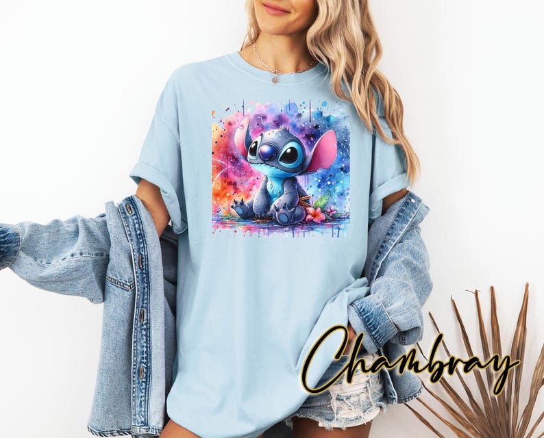 May include: Pale blue t-shirt with a watercolour illustration of the Disney character Stitch. The artwork depicts Stitch in a seated position against a backdrop of pink, blue, and purple colours. The t-shirt is made of a soft, comfortable material.