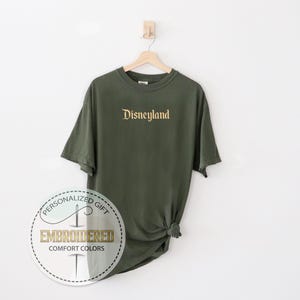 Pu&ograve; includere: T-shirt verde oliva con la scritta "Disneyland" ricamata in oro sul petto. La maglietta &egrave; appesa a una gruccia di legno su sfondo bianco. Un logo circolare con le parole "Embroidered Comfort Colors" &egrave; in basso.