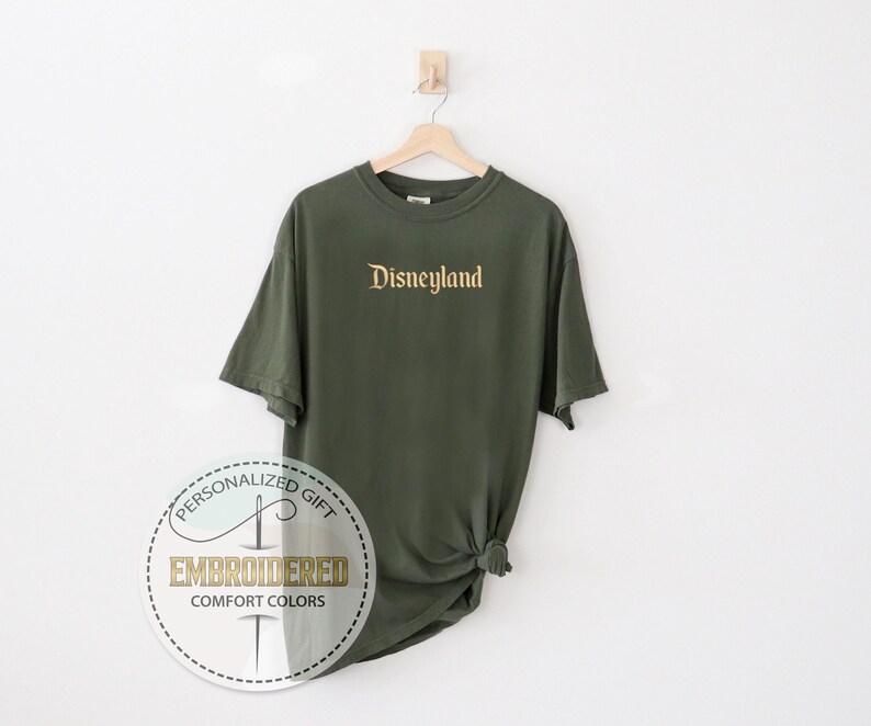 Pu&ograve; includere: T-shirt verde oliva con la scritta "Disneyland" ricamata in oro sul petto. La maglietta &egrave; appesa a una gruccia di legno su sfondo bianco. Un logo circolare con le parole "Embroidered Comfort Colors" &egrave; in basso.