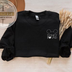 May include: Black crewneck sweatshirt with the text "DISNEY BOUND" and a Mickey Mouse head outline. The sweatshirt has long sleeves and a simple design. The image also includes a decorative element with the text "EMBROIDERED SWEATSHIRT & HOODIE".