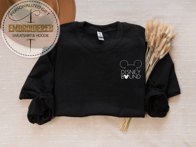 May include: Black crewneck sweatshirt with the text "DISNEY BOUND" and a Mickey Mouse head outline. The sweatshirt has long sleeves and a simple design. The image also includes a decorative element with the text "EMBROIDERED SWEATSHIRT & HOODIE".