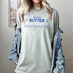 May include: A pale green t-shirt featuring the words "SALTED BUTTER" in blue, and a measuring guide. The shirt also has the text "NET WT. 4 OZ (113 g)" and "GRADE AA". A denim jacket is worn over the t-shirt.