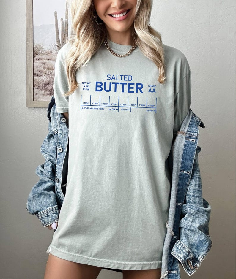 May include: A pale green t-shirt featuring the words "SALTED BUTTER" in blue, and a measuring guide. The shirt also has the text "NET WT. 4 OZ (113 g)" and "GRADE AA". A denim jacket is worn over the t-shirt.