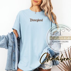 Pu&ograve; includere: T-shirt azzurra con la scritta "Disneyland" ricamata in marrone. La maglietta ha una vestibilit&agrave; ampia e fa parte di una collezione "Comfort Colors". Una giacca di jeans &egrave; drappeggiata sulle spalle.
