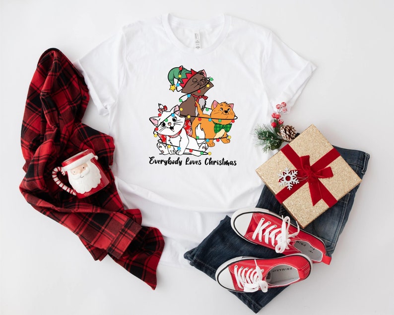 May include: White t-shirt with a Christmas-themed design featuring three cats wearing Santa hats and surrounded by Christmas lights. The text "Everybody Loves Christmas" is printed below the cats.
