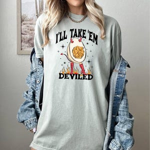以下が含まれることがあります： 薄い緑色のTシャツに、角と赤いブーツを履いた悪魔の卵のグラフィックがプリントされています。Tシャツには「I'll take 'em deviled」という文字がプリントされています。