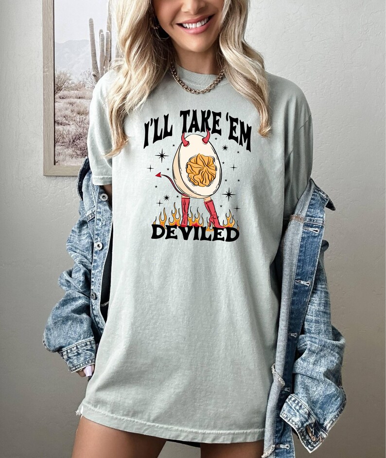 以下が含まれることがあります： 薄い緑色のTシャツに、角と赤いブーツを履いた悪魔の卵のグラフィックがプリントされています。Tシャツには「I'll take 'em deviled」という文字がプリントされています。