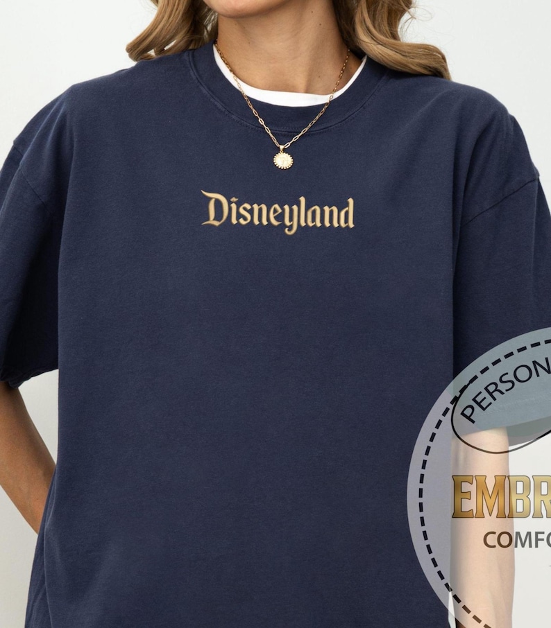 Pu&ograve; includere: Felpa blu navy girocollo con la scritta "Disneyland" ricamata in oro sul petto. La felpa ha maniche corte e una maglietta bianca &egrave; visibile allo scollo. Si indossa una collana d'oro con un ciondolo.