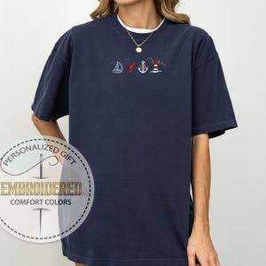 May include: Navy blue t-shirt with embroidered nautical designs, including a sailboat, anchor, and lighthouse. The shirt has a white crew neck collar and a gold necklace. The shirt is labeled "Embroidered Comfort Colors".