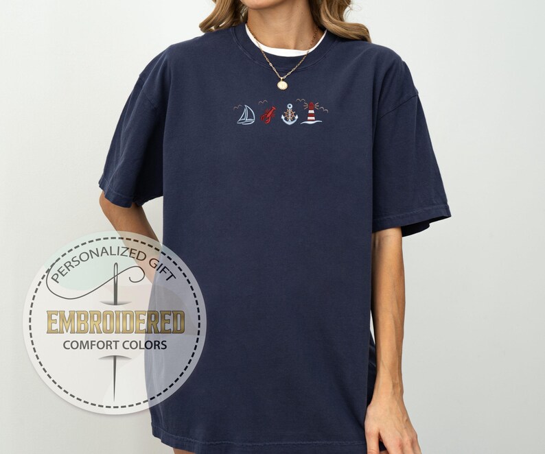 May include: Navy blue t-shirt with embroidered nautical designs, including a sailboat, anchor, and lighthouse. The shirt has a white crew neck collar and a gold necklace. The shirt is labeled "Embroidered Comfort Colors".