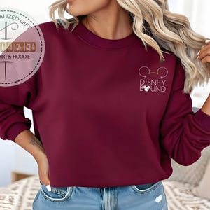 May include: Burgundy sweatshirt with the text "Disney Bound" and a Mickey Mouse head outline. The sweatshirt has a crew neck and long sleeves. The image also includes a logo that says "Embroidered Sweatshirt & Hoodie".