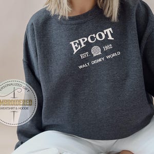 May include: A dark grey EPCOT sweatshirt featuring white text that reads "EPCOT", "EST. 1982", and "WALT DISNEY WORLD". The design includes a globe graphic. Part of the Embroidered Sweatshirt & Hoodie collection.