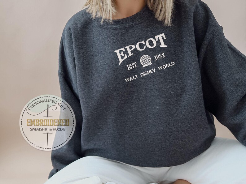 May include: A dark grey EPCOT sweatshirt featuring white text that reads "EPCOT", "EST. 1982", and "WALT DISNEY WORLD". The design includes a globe graphic. Part of the Embroidered Sweatshirt & Hoodie collection.