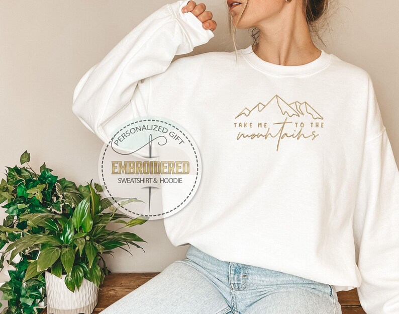 K&ouml;nnte beinhalten: Wei&szlig;es Sweatshirt mit goldener Berggrafik und dem Text "Take me to the mountains". Das Sweatshirt hat lange &Auml;rmel. Ein kreisf&ouml;rmiges Logo lautet "Embroidered Sweatshirt & Hoodie".