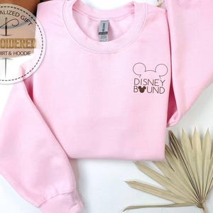 May include: A light pink sweatshirt with the text "Disney Bound" and a Mickey Mouse head graphic in brown. The sweatshirt has long sleeves and a crew neck. The image also includes a decorative dried plant.