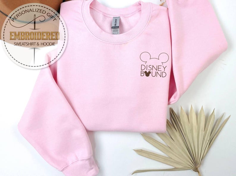 May include: A light pink sweatshirt with the text "Disney Bound" and a Mickey Mouse head graphic in brown. The sweatshirt has long sleeves and a crew neck. The image also includes a decorative dried plant.