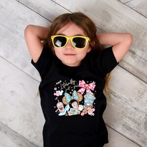 May include: A young person wearing a black t-shirt with the text "Literally just a girl" and colorful graphics of bows, desserts, and a high heel. The person is also wearing yellow sunglasses.