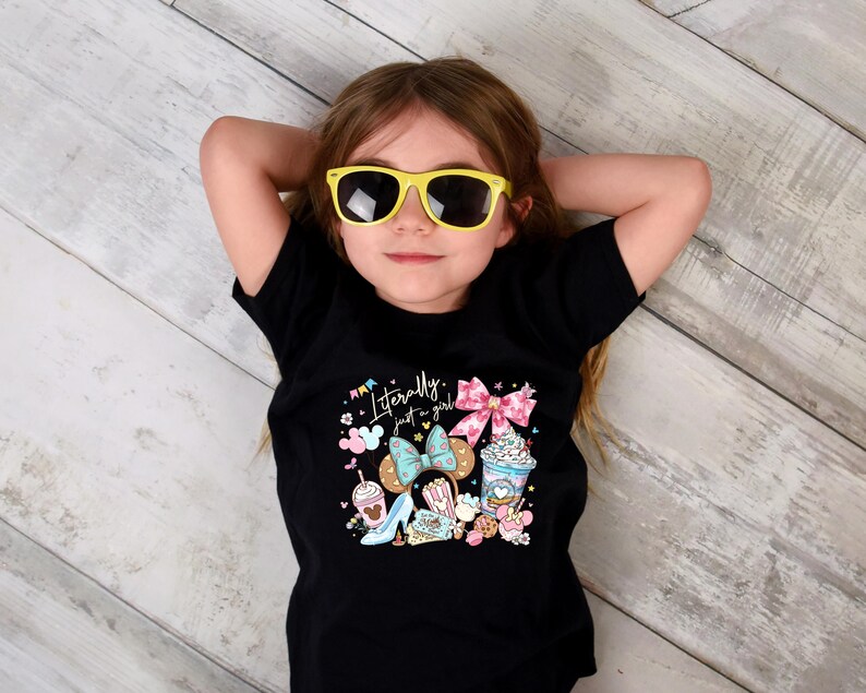 May include: A young person wearing a black t-shirt with the text "Literally just a girl" and colorful graphics of bows, desserts, and a high heel. The person is also wearing yellow sunglasses.