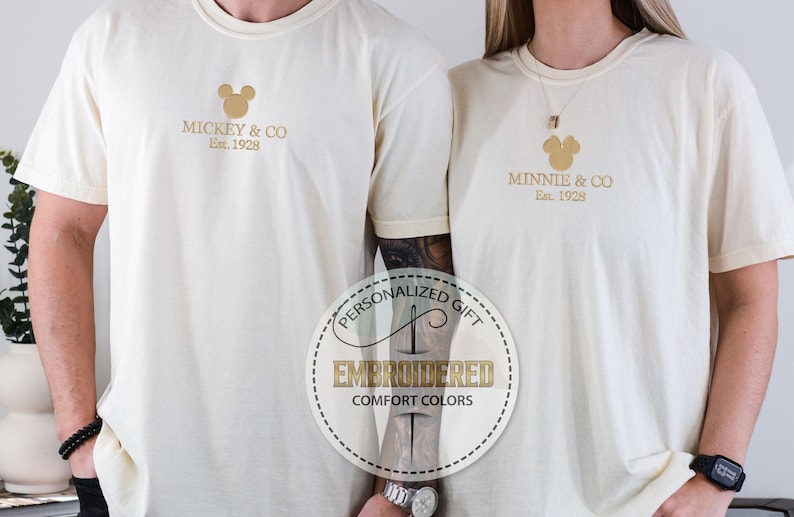 May include: Cream-colored t-shirts with gold embroidered designs. One shirt features "Mickey & Co. Est. 1928," and the other "Minnie & Co. Est. 1928." A circular graphic reads "Personalized Gift" and "Embroidered Comfort Colors."