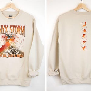 May include: A beige sweatshirt with a graphic print of a mountain, a dragon, and the text "ONYX STORM CREATIVE THE FUTURE I WILL SAVE HIM". The back of the sweatshirt has a vertical row of five dragon silhouettes.