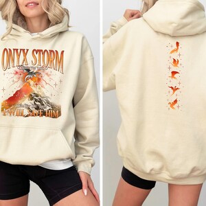 May include: A cream-colored hoodie with a graphic print of a dragon and the text "ONYX STORM" and "I WILL SAVE HIM". The back of the hoodie has a vertical line of five orange dragon silhouettes.
