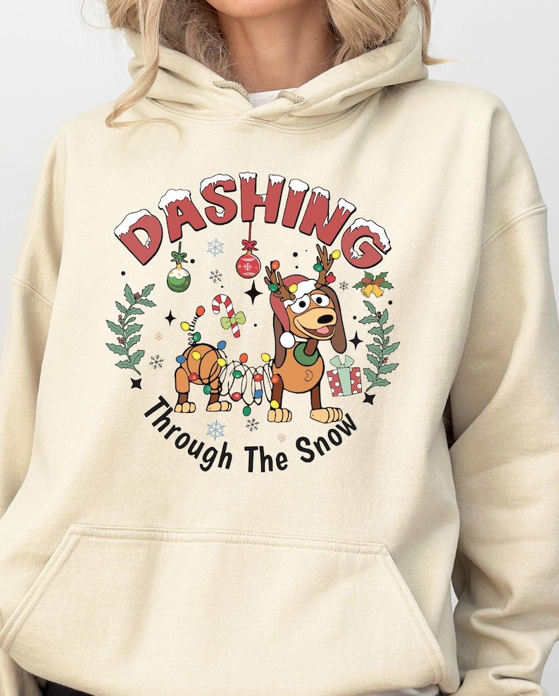 K&ouml;nnte beinhalten: Beige Kapuzenpullover mit einem weihnachtlichen Motiv. Das Design zeigt einen Cartoon-Hund mit Geweih und Lichterketten, mit dem Text "Dashing Through The Snow". Festliche Ornamente und eine Geschenkbox sind ebenfalls vorhanden.