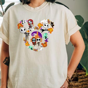 May include: A white t-shirt with a Mickey Mouse silhouette design featuring characters from the Nightmare Before Christmas movie. The design includes Jack Skellington, Sally, and other characters from the film.