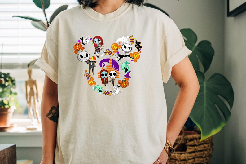 May include: A white t-shirt with a Mickey Mouse silhouette design featuring characters from the Nightmare Before Christmas movie. The design includes Jack Skellington, Sally, and other characters from the film.