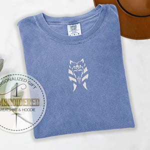 May include: A blue t-shirt with a white embroidered design of a Star Wars character's symbol. The text "Personalized Gift" and "Embroidered Sweatshirt & Hoodie" is on a white circle sticker.