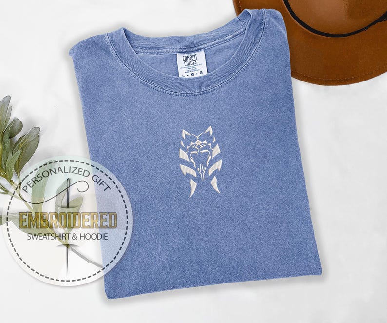 May include: A blue t-shirt with a white embroidered design of a Star Wars character's symbol. The text "Personalized Gift" and "Embroidered Sweatshirt & Hoodie" is on a white circle sticker.