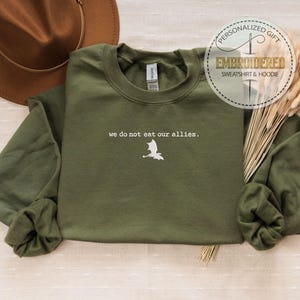 May include: A green long-sleeved sweatshirt with the text "we do not eat our allies." and a white dragon silhouette embroidered on the front.