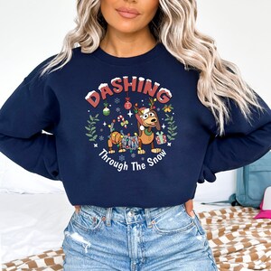 K&ouml;nnte beinhalten: Marineblaues Sweatshirt mit einem weihnachtlichen Design. Die Grafik zeigt einen Dackel mit Geweih, umgeben von festlichen Dekorationen. Der Text lautet "Dashing Through The Snow", ideal f&uuml;r die Weihnachtszeit.