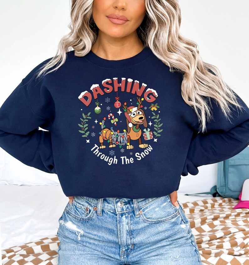 K&ouml;nnte beinhalten: Marineblaues Sweatshirt mit einem weihnachtlichen Design. Die Grafik zeigt einen Dackel mit Geweih, umgeben von festlichen Dekorationen. Der Text lautet "Dashing Through The Snow", ideal f&uuml;r die Weihnachtszeit.