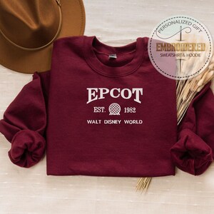 May include: A maroon sweatshirt with the text "EPCOT EST. 1982 WALT DISNEY WORLD" in white. The sweatshirt is displayed with a brown hat and decorative wheat stalks. A circular graphic reads "PERSONALIZED GIFT EMBROIDERED SWEATSHIRT & HOODIE".