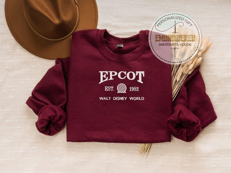 May include: A maroon sweatshirt with the text "EPCOT EST. 1982 WALT DISNEY WORLD" in white. The sweatshirt is displayed with a brown hat and decorative wheat stalks. A circular graphic reads "PERSONALIZED GIFT EMBROIDERED SWEATSHIRT & HOODIE".