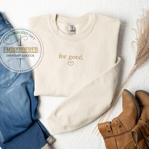 May include: A cream-colored sweatshirt with the text "for good." embroidered in gold with a small heart symbol. The sweatshirt is laid out on a white surface with a pair of brown boots and a pair of blue jeans.