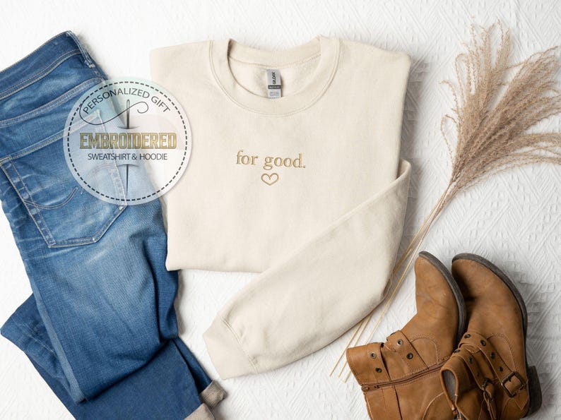 May include: A cream-colored sweatshirt with the text "for good." embroidered in gold with a small heart symbol. The sweatshirt is laid out on a white surface with a pair of brown boots and a pair of blue jeans.