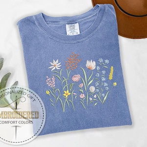May include: A light blue t-shirt with an embroidered floral design. The flowers are in shades of pink, yellow, and white. The shirt is made of a soft, comfortable fabric.