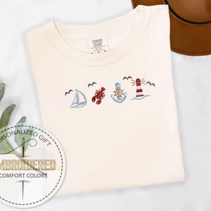 May include: Cream-colored Comfort Colors t-shirt with embroidered nautical designs: sailboat, lobster, anchor, and lighthouse. The shirt also has the text "EMBROIDERED COMFORT COLORS" and "PERSONALIZED GIFT" in a circular design.