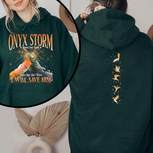 May include: A dark green hoodie with a graphic print of a mountain and the text "ONYX STORM - BRAVE THE DARK - I WILL NOT DIE TODAY - I WILL SAVE HIM". The back of the hoodie has a vertical row of five gold dragon silhouettes.