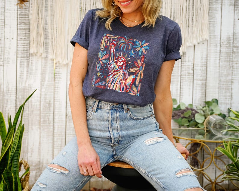 May include: A navy blue t-shirt with a colorful graphic of the Statue of Liberty surrounded by flowers and fireworks. The graphic is framed by a white rectangle.