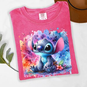 May include: A pink Comfort Colors t-shirt with a watercolour illustration of Stitch from Lilo & Stitch. The design features colourful splashes and a floral detail. The shirt is a size L-G-G.