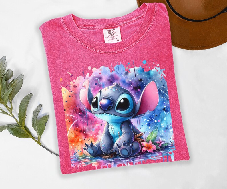 May include: A pink Comfort Colors t-shirt with a watercolour illustration of Stitch from Lilo & Stitch. The design features colourful splashes and a floral detail. The shirt is a size L-G-G.