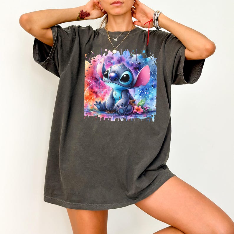 May include: Dark grey t-shirt with a watercolour illustration of the character Stitch from Lilo & Stitch. The artwork shows Stitch in blue with pink ears, against a vibrant, colourful background. The shirt is a casual, oversized style.